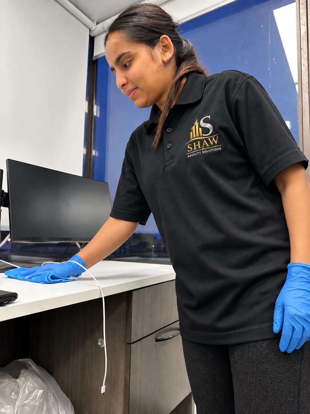 Campbell Shaw team member in a branded polo and nitrile gloves wiping down a desk beside a monitor in an office