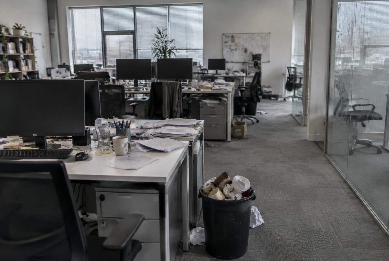 Open-plan office floor with cluttered desks, dust, scattered paper, an overflowing waste bin and soiled carpet before cleaning